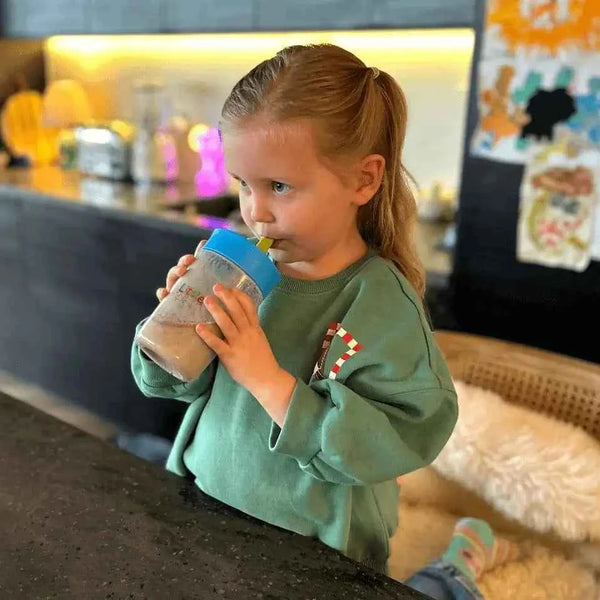 Lifestyle photo of a child drinking LittleVitz Kids Super Greens chocolate flavour from a reusable LittleVitz cup in a home kitchen. The image demonstrates how the daily greens and superfoods drink fits into a child's everyday routine, providing a convenient and enjoyable way for children aged 3+ to get vitamins, minerals and plant based nutrition.
